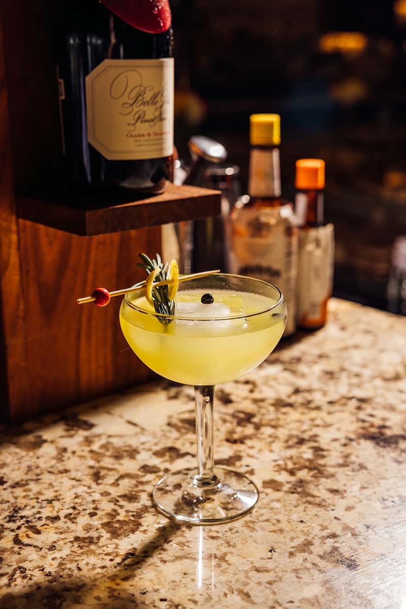 Elegant craft cocktail with a vibrant yellow hue served in a coupe glass, garnished with a rosemary sprig, lemon twist, and a single juniper berry, set on a granite bar counter with bottles of bitters and wine in the background—perfect for upscale bar or cocktail lounge ambiance.