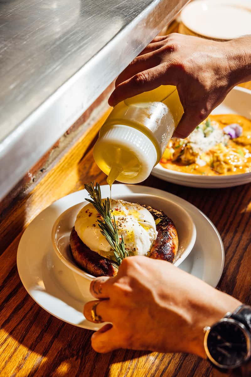 Close-up of a chef drizzling olive oil over grilled Italian sausage topped with creamy burrata and garnished with rosemary, highlighting final plating touches in a rustic Italian kitchen—perfect for showcasing rich, handcrafted appetizers with authentic flavor.
