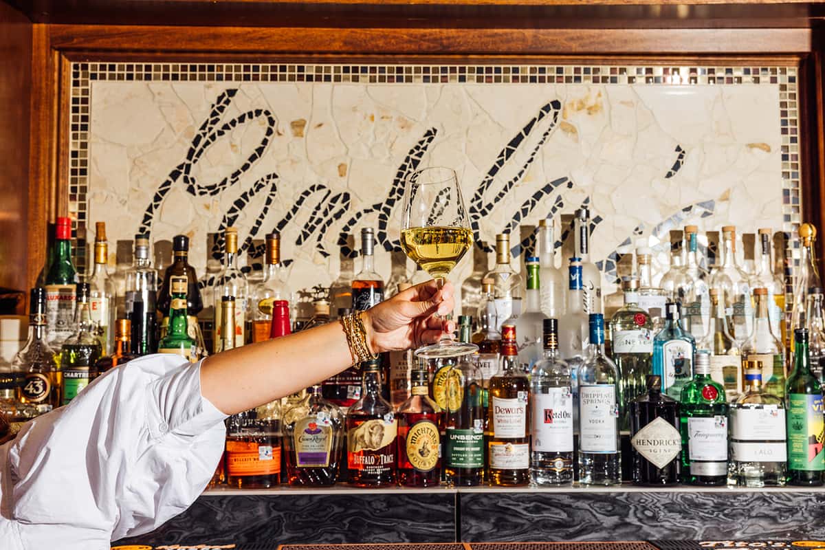 Woman raising a glass of white wine in front of a fully stocked bar featuring premium liquor bottles and a mosaic tile backdrop that reads "Punky's"—ideal for highlighting upscale bar offerings and vibrant cocktail lounge atmosphere.