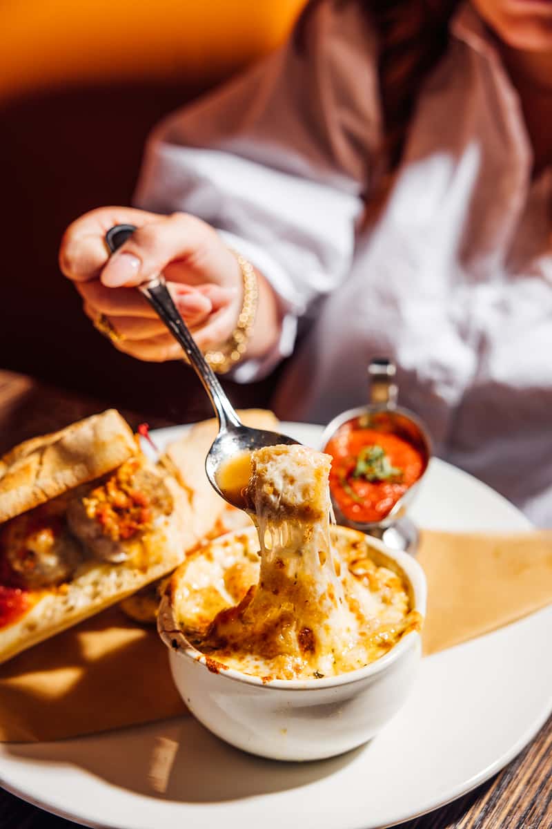 Close-up of a cheesy baked pasta dish being lifted with a spoon, showcasing gooey melted mozzarella, served alongside a hearty meatball sub and marinara dipping sauce—ideal for highlighting indulgent Italian-American comfort food.