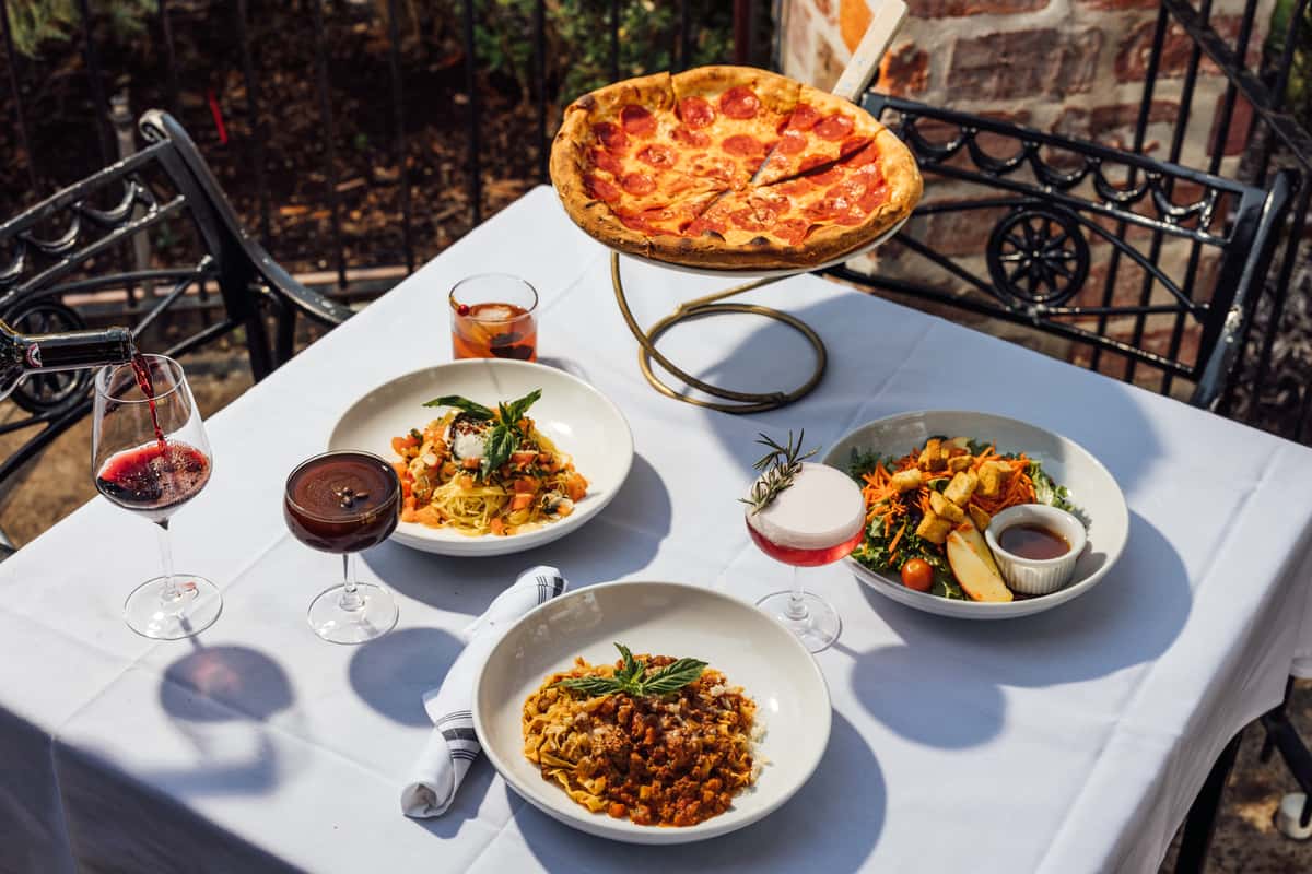 Outdoor Italian dining table set with a pepperoni pizza on a raised stand, fresh garden salad, creamy burrata pasta, rich Bolognese tagliatelle, and a variety of cocktails and red wine—perfect for highlighting authentic Italian cuisine and a vibrant alfresco restaurant experience.