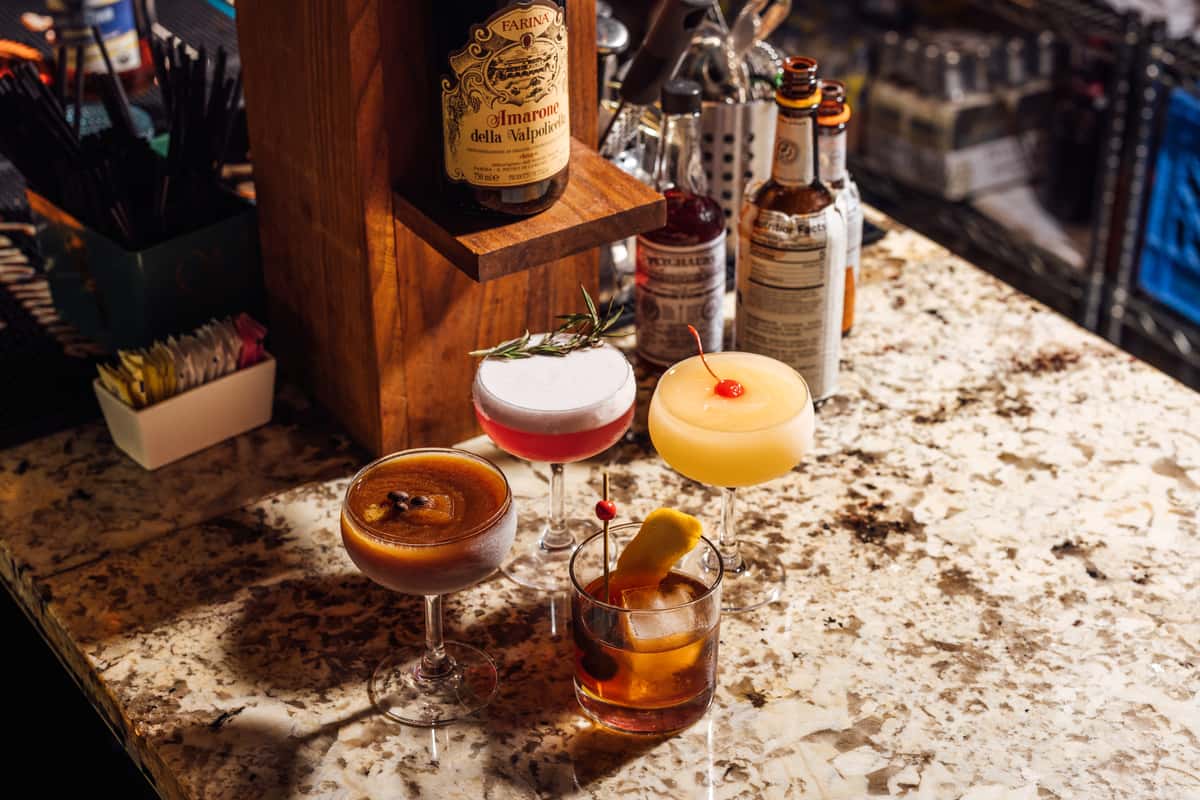 A variety of colorful craft cocktails displayed on a marble bar counter, including a frozen drink garnished with a cherry, a frothy pink cocktail with a rosemary sprig, a classic Old Fashioned, and an espresso martini, with a bottle of Amarone wine and bitters in the background.