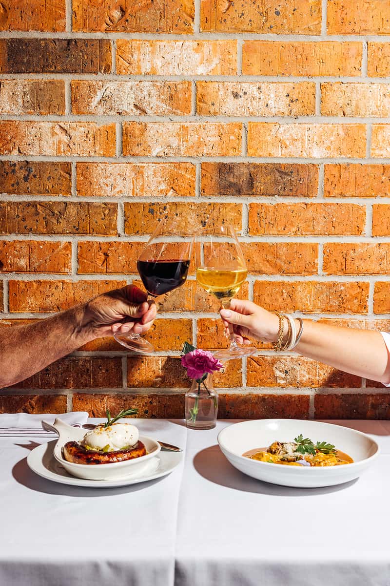 Two diners toasting with glasses of red and white wine over a white tablecloth setting, with Italian dishes including grilled sausage with burrata and creamy stuffed pasta in the foreground, set against a rustic brick wall—perfect for capturing romantic dining or celebratory moments at an Italian restaurant.