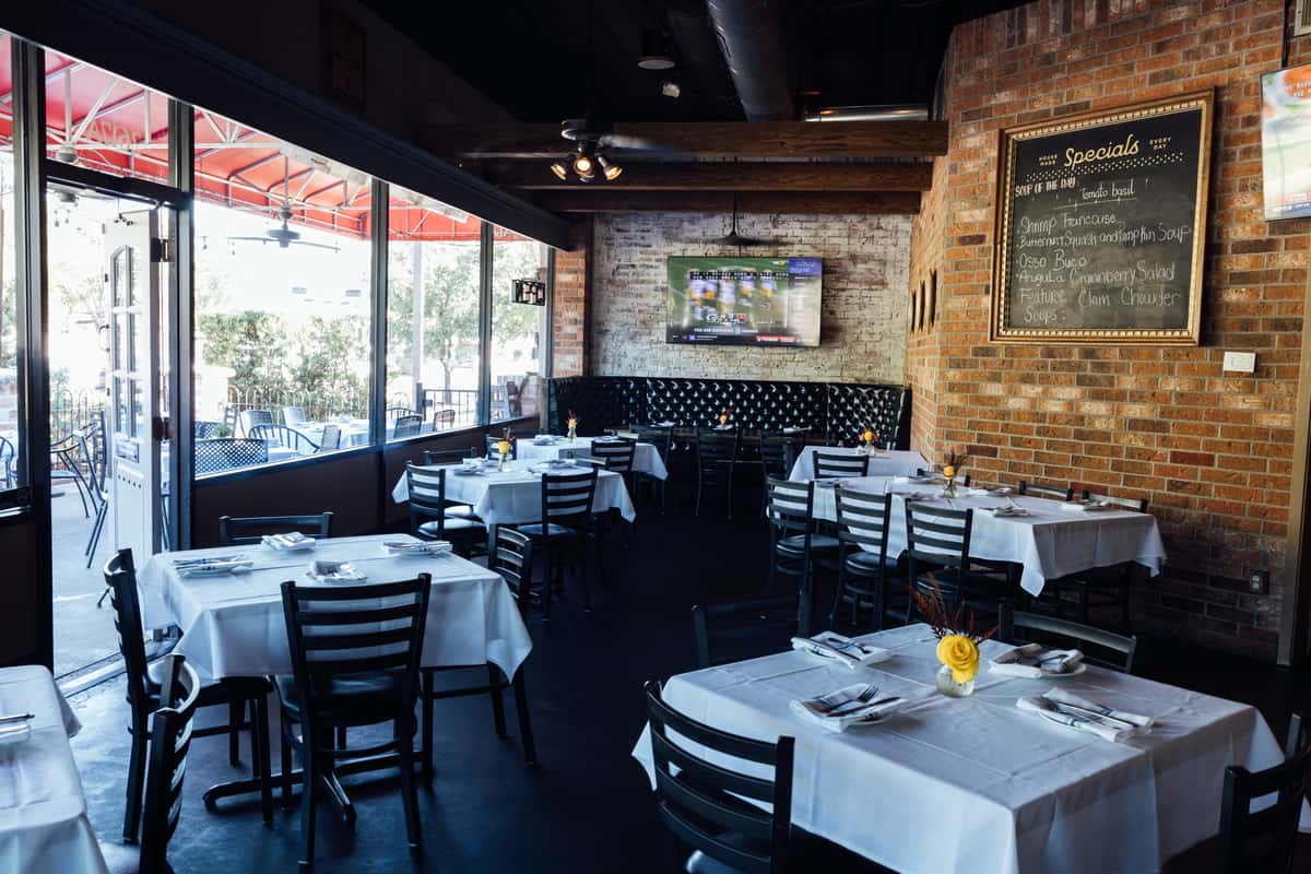 Cozy Italian restaurant dining room with black chairs, white tablecloths, and a rustic brick interior, featuring a large chalkboard specials menu, flat-screen TV, and natural light streaming through large windows—ideal for showcasing a welcoming and casual yet refined dining atmosphere.