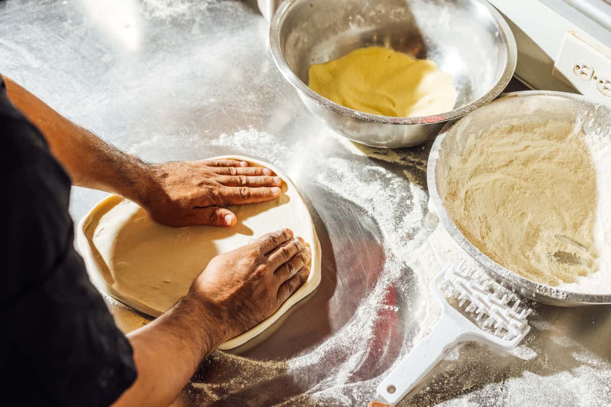 Chef shaping fresh pizza dough by hand on a floured stainless steel prep table, surrounded by bowls of flour and dough—highlighting traditional pizza-making techniques and artisanal preparation in an authentic Italian kitchen.