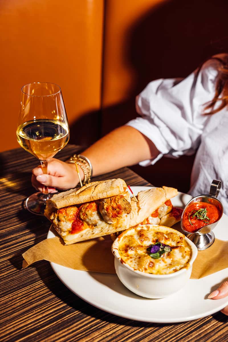 Hearty Italian meal featuring a toasted meatball sub topped with marinara and melted cheese, served alongside a baked dish of cheesy pasta or lasagna and a side of tomato basil sauce, paired with a glass of chilled white wine—ideal for showcasing cozy Italian-American comfort food.