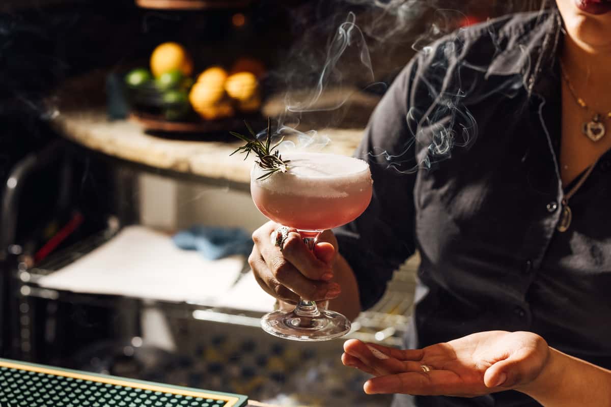 Bartender presenting a smoky pink craft cocktail topped with foam and garnished with a smoldering rosemary sprig, capturing a dramatic moment of mixology at a modern Italian bar—perfect for highlighting signature cocktails and elevated drink presentation.