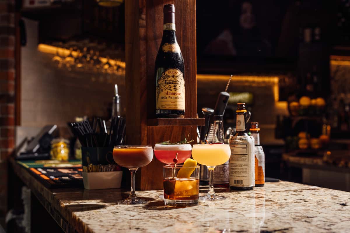 Lineup of vibrant craft cocktails on a granite bar counter, including whiskey and citrus-based drinks garnished with rosemary, cherries, and lemon peel, set beneath a bottle of Amarone della Valpolicella in a dimly lit Italian bar—ideal for promoting artisanal cocktails and upscale bar ambiance.