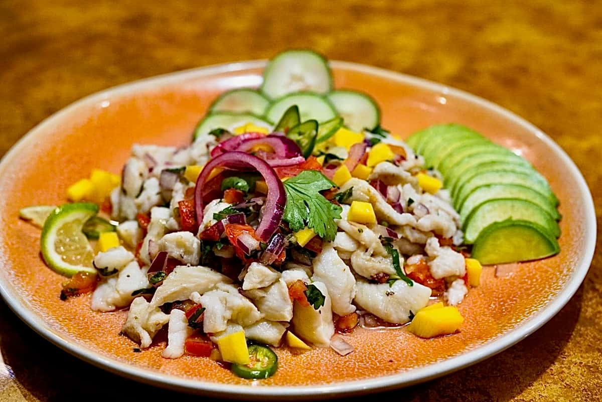 Fresh fish ceviche with mango, red onion, cilantro, and jalapeño, garnished with cucumber and lime slices on an orange plate.