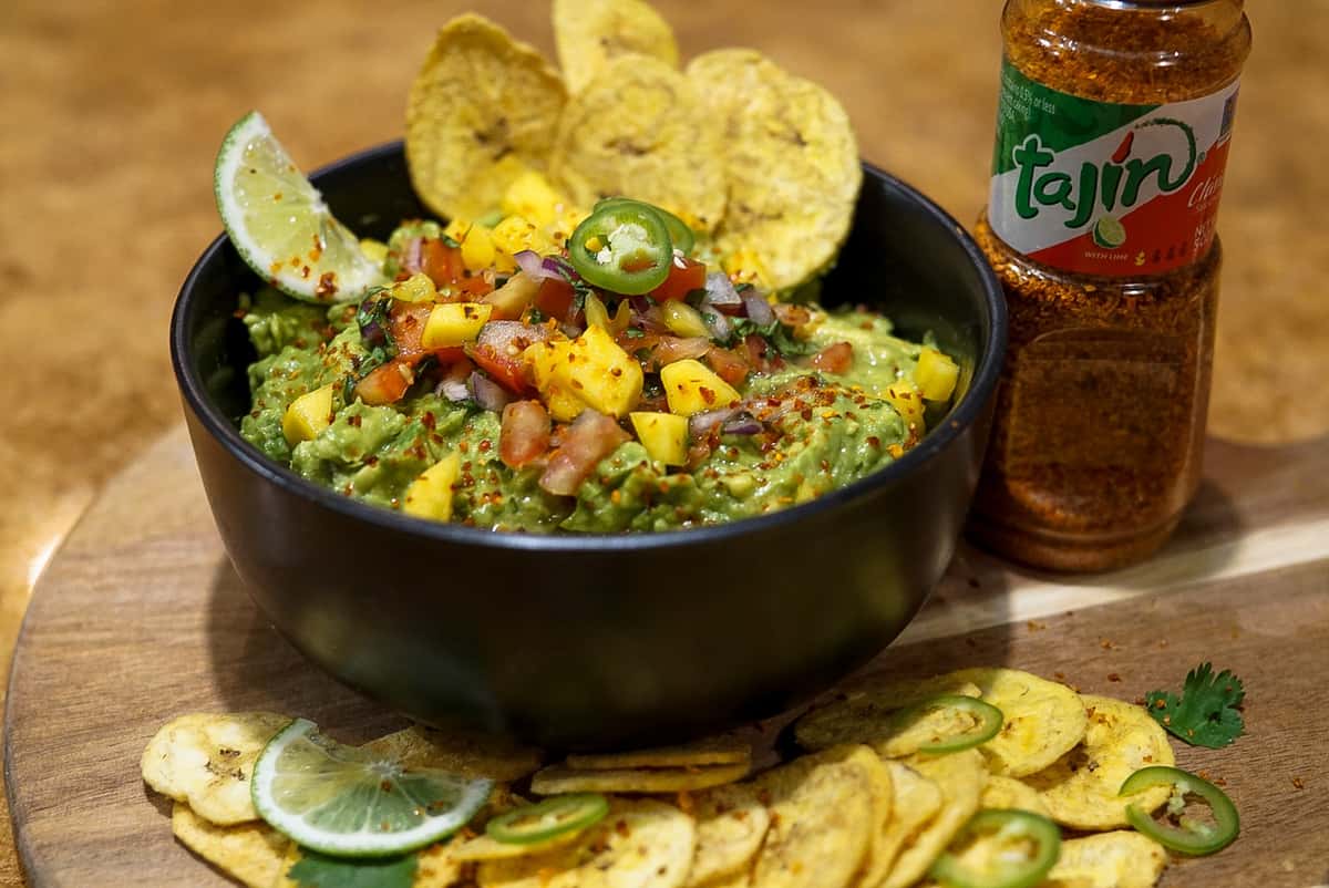 Tropical guacamole topped with mango salsa, diced tomatoes, red onion, jalapeño, and Tajín seasoning, served in a black bowl with crispy plantain chips, lime wedges, and a bottle of Tajín at La Delicia Mexican Brunch & Cantina.