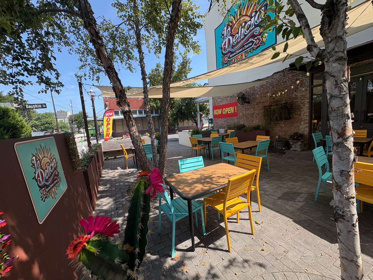 Colorful outdoor patio at La Delicia with yellow and teal seating, shaded by sails and surrounded by trees and vibrant signage.