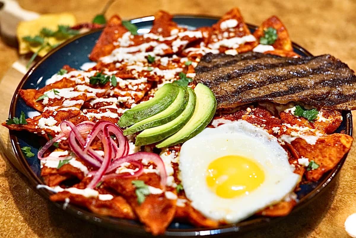 Plate of traditional Mexican chilaquiles rojos topped with a sunny-side-up egg, sliced avocado, grilled steak, pickled red onions, fresh cilantro, and a drizzle of crema, served at La Delicia Mexican Brunch & Cantina.