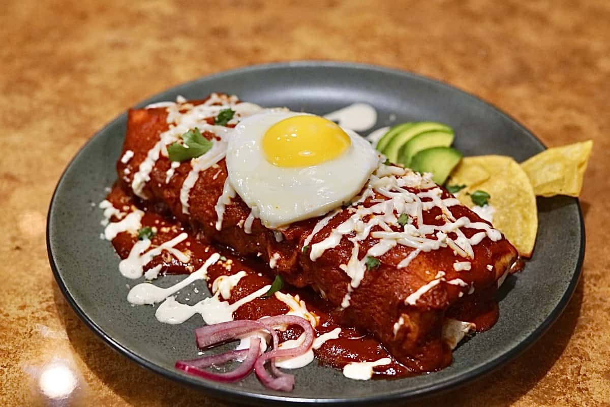 Smothered enchiladas topped with a sunny-side-up egg, drizzled with crema, and served with avocado slices and tortilla chips.