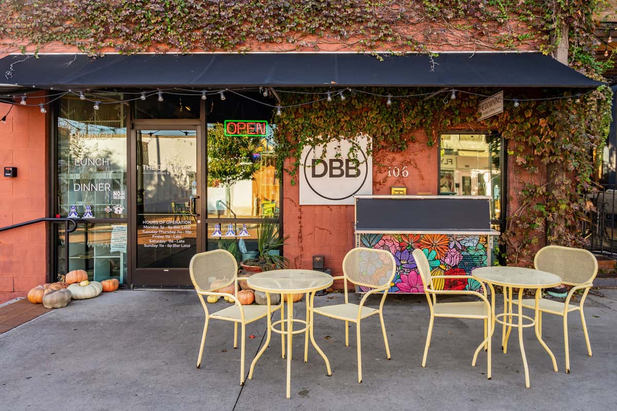 Exterior view of The Downing Bottles and Bites showing yellow metal bistro seating, a black awning, and an ivy-covered terra cotta brick wall.