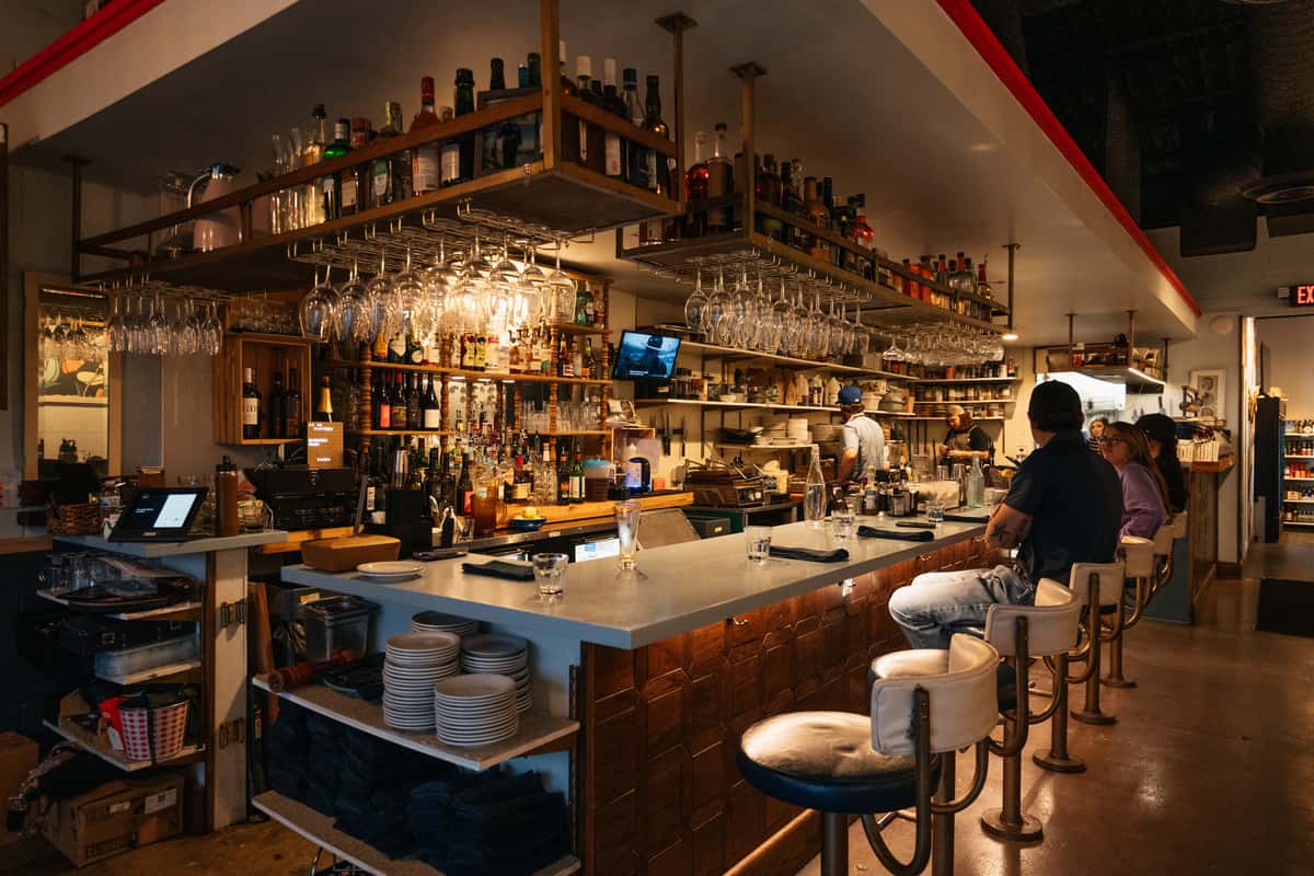 Warmly lit bar interior at The Downing Bottles & Bites featuring stacked liquor bottles, hanging glasses, and seated patrons at the counter.