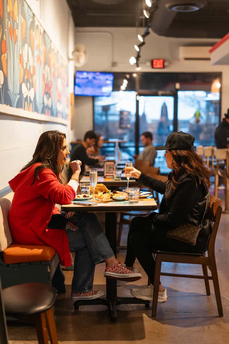 Interior view of The Downing Bottles & Bites showing two women, one in red, toasting with champagne and sharing a cheese board next to a colorful floral wall mural.