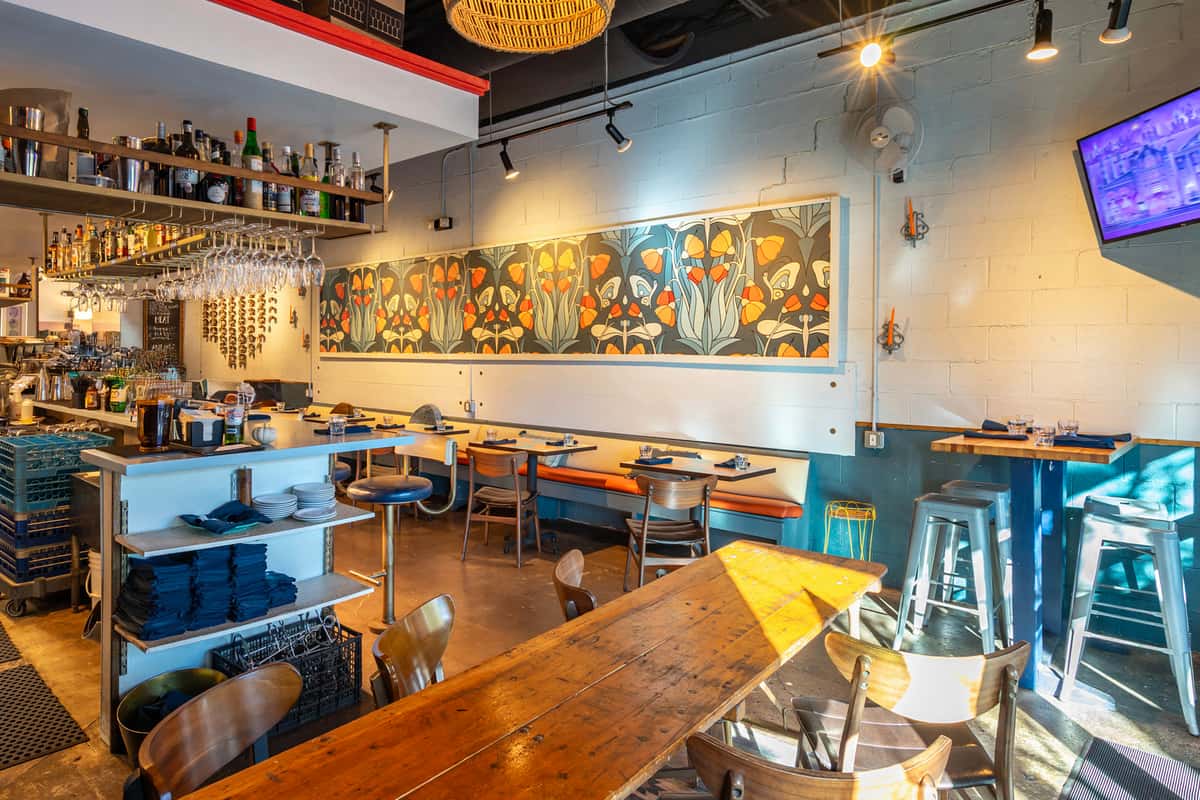 The Downing Bottles & Bites restaurant interior with communal wood table, gray walls, and bright orange floral mural over booth seating.