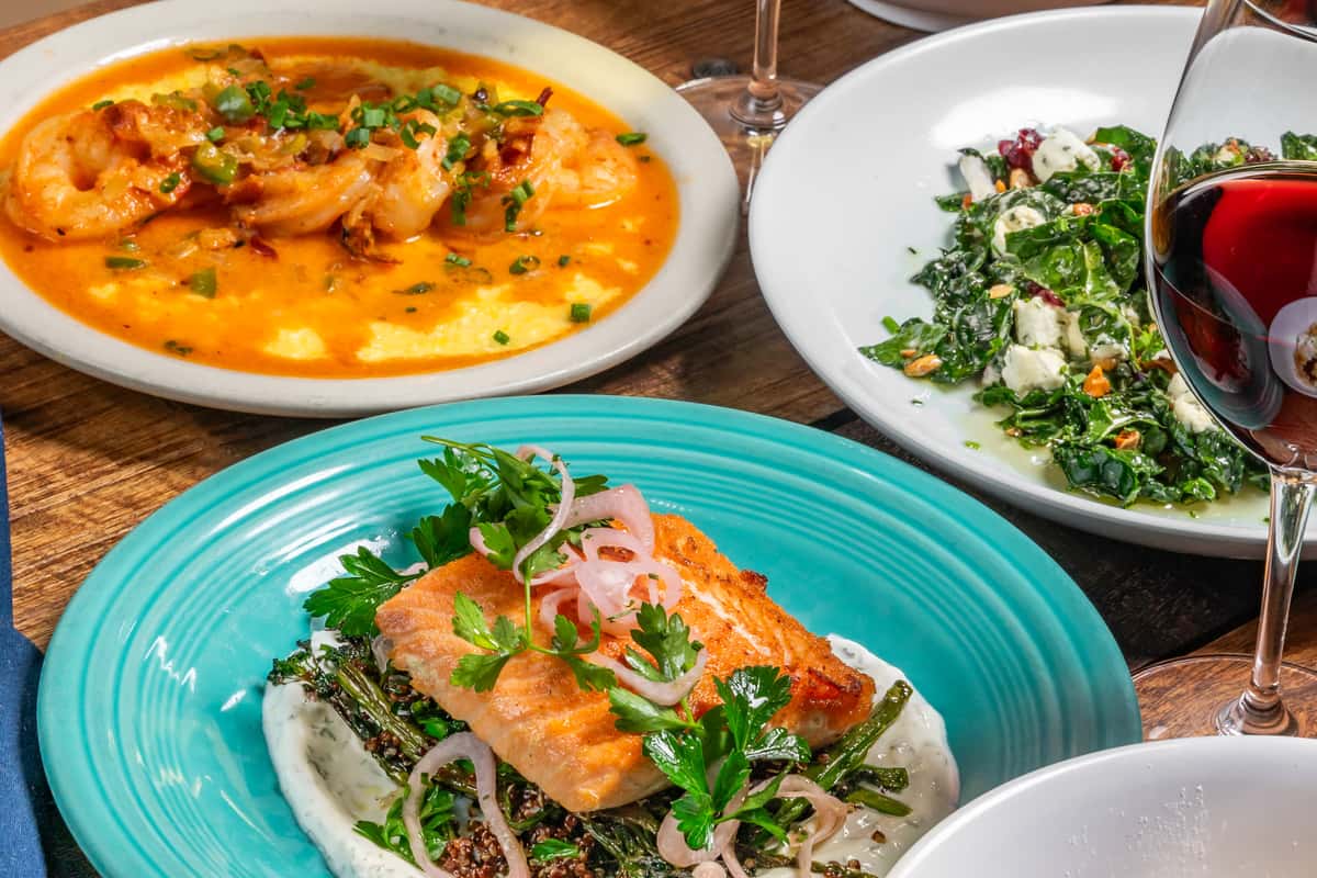 Colorful close-up of plated dishes including grilled salmon, shrimp and grits, and kale salad with red wine at The Downing Bottles and Bites.