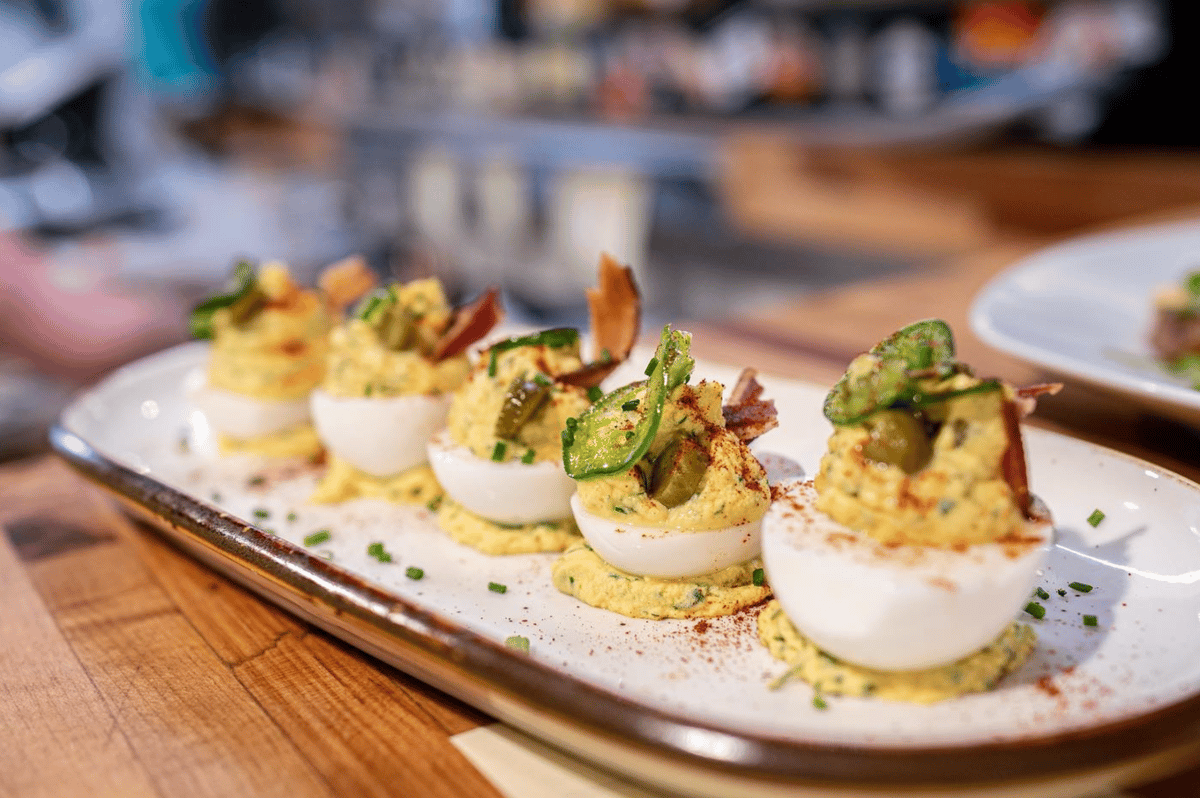 Five gourmet deviled eggs garnished with bacon and pickles, served on a white plate at The Downing Bottles and Bites.