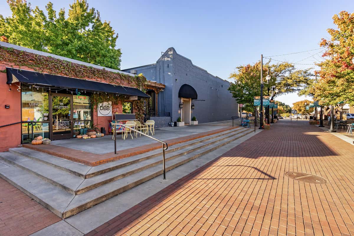 The Downing Bottles and Bites restaurant exterior features a vine-covered red facade, outdoor seating, and concrete steps leading to a sunlit red brick pedestrian street.