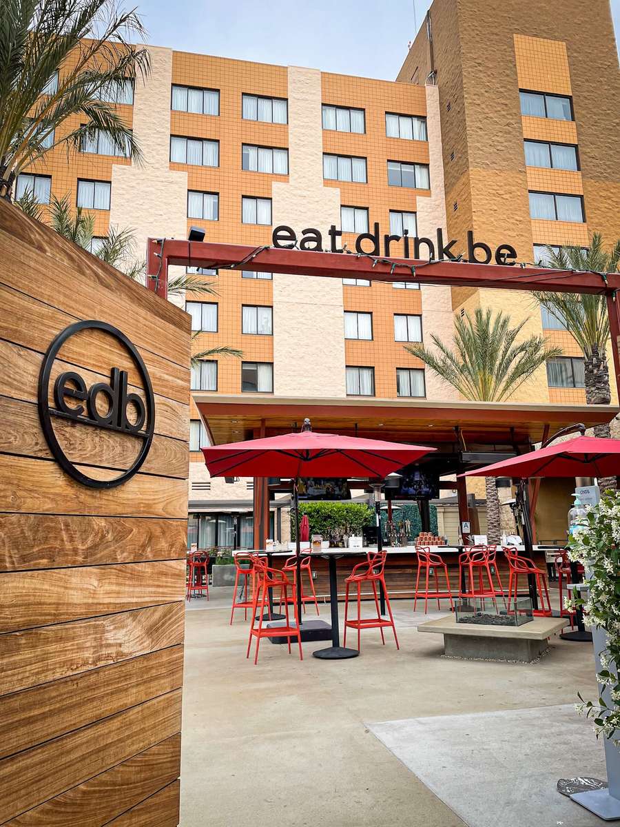 Outdoor patio bar at edb (eat. drink. be.) featuring red umbrellas, modern seating, palm trees, and an open-air lounge in front of the hotel.