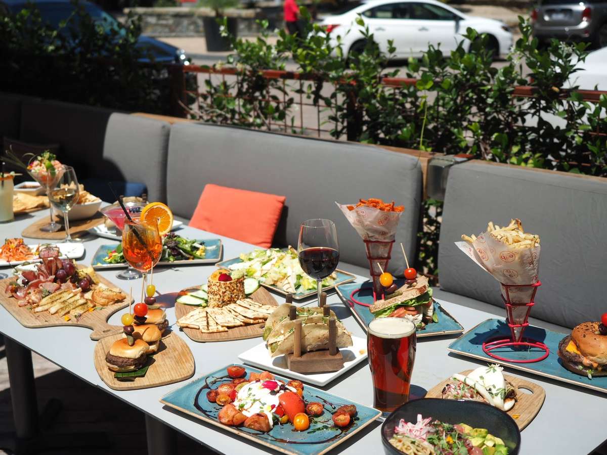Outdoor patio dining with gourmet burgers, shareable appetizers, craft cocktails, beer, and fresh salads served on a sunny restaurant terrace.
