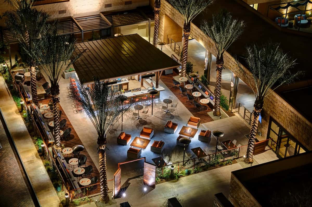 Aerial nighttime view of the edb (eat. drink. be.) outdoor patio featuring a modern open-air bar, fire pit lounge seating, palm trees, and stylish dining areas.