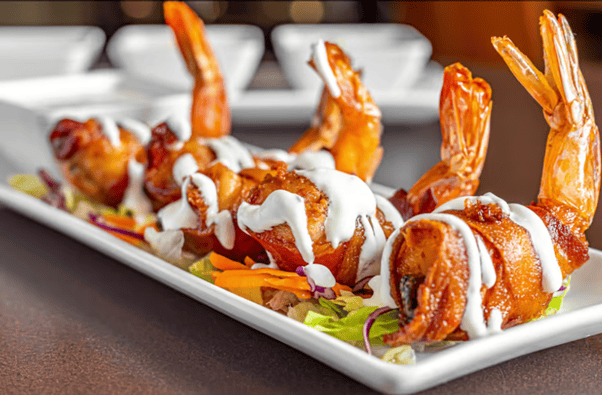Mesero’s bacon-wrapped shrimp drizzled with creamy white sauce, served on a white rectangular plate.