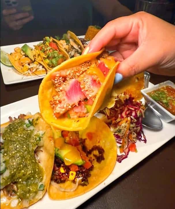 Mesero tacos with seared tuna, fresh toppings, and colorful sides, with a guest holding one taco.
