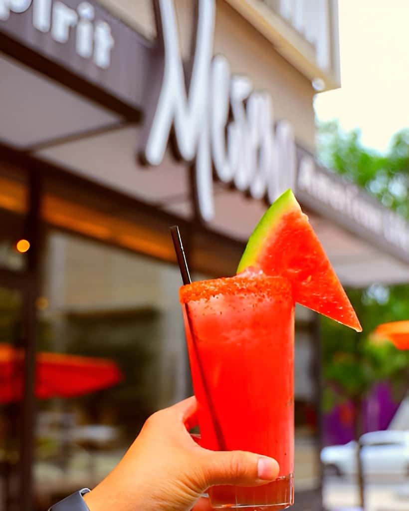 Hand holding a refreshing watermelon cocktail in a transparent glass with a watermelon wedge and straw in front of the Mesero restaurant exterior.