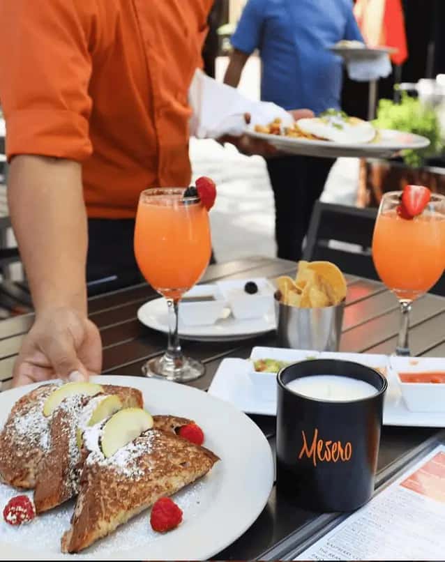 Brunch at Mesero with French toast, orange cocktails, chips, salsa, and servers in bright uniforms at an outdoor table.