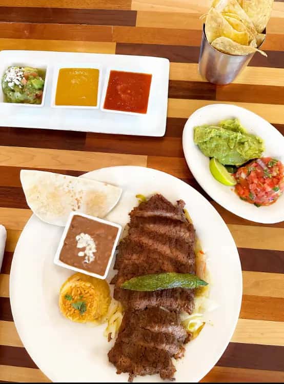 Mesero steak plate with rice, beans, tortillas, guacamole, salsa trio, and chips served on a wooden table.