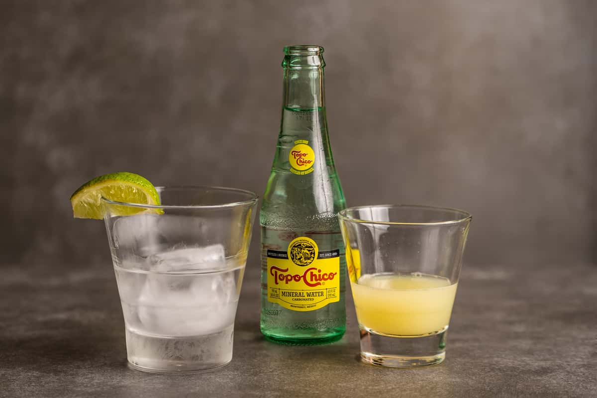 Tequila cocktail setup by Mesero with mineral water bottle, lime garnish, and ice-filled glass.