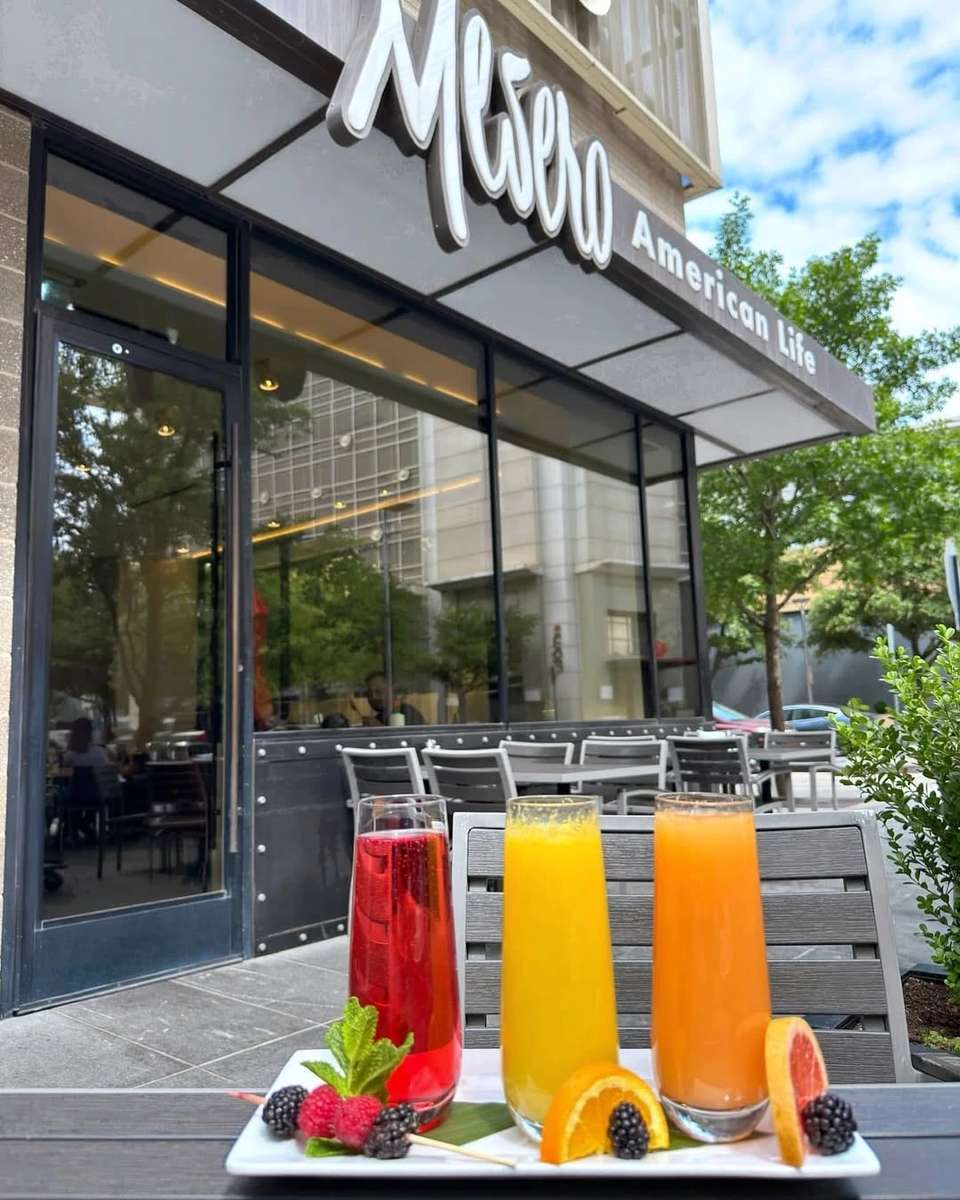 Three colorful drinks (red, yellow, orange) with fruit garnishes on an outdoor patio table in front of Mesero.