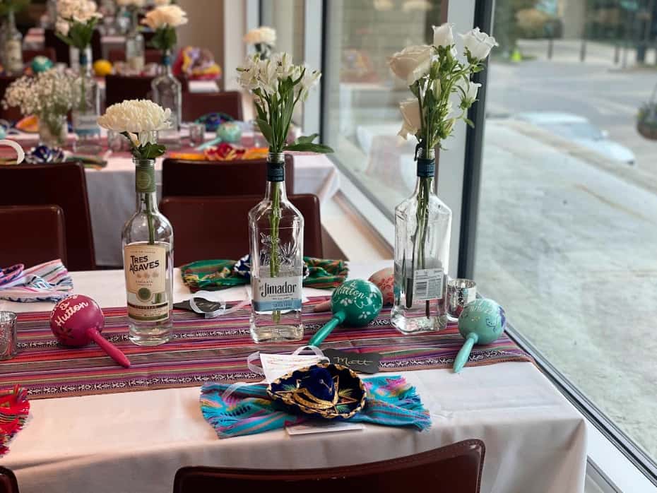 Mesero table decor with tequila bottle vases, white flowers, and colorful fiesta accents by window.