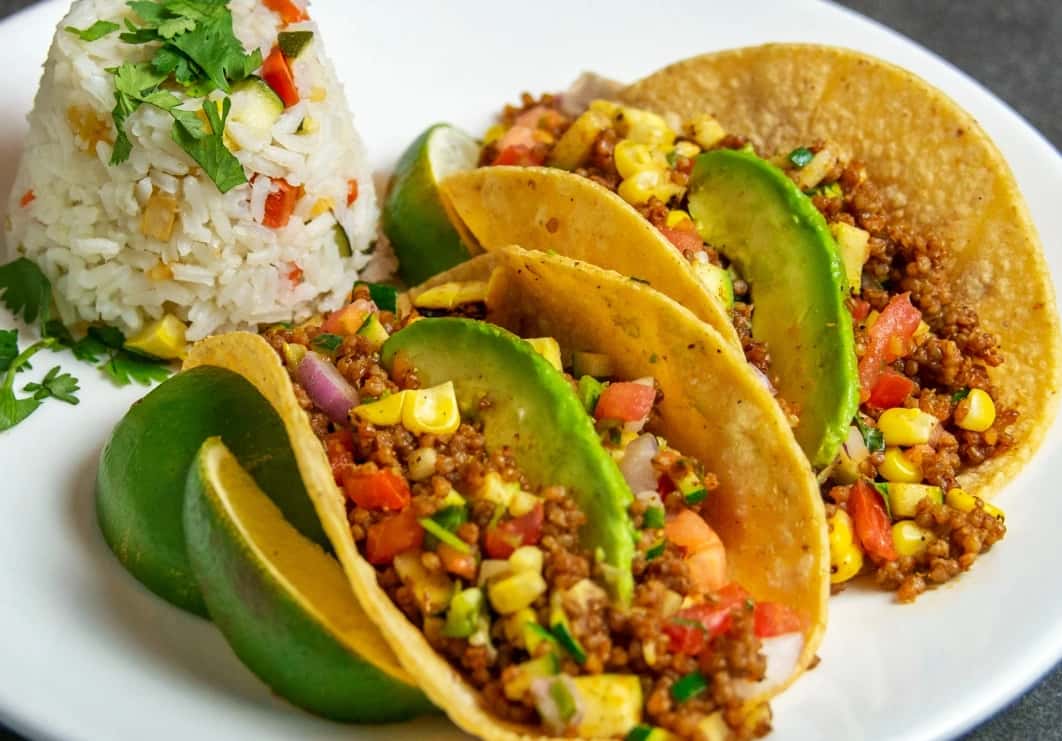 Mesero quinoa tacos topped with avocado and fresh corn salsa, served with seasoned rice on a vibrant plate.