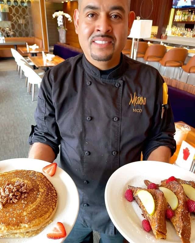 A Mesero chef holding a plate of pancakes topped with nuts and French toast with fruits.