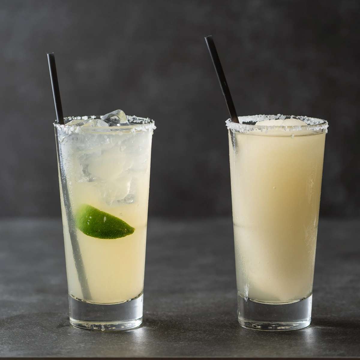 Two salted-rim margarita glasses, one classic and one frozen, with lime and ice on a dark background at Mesero.