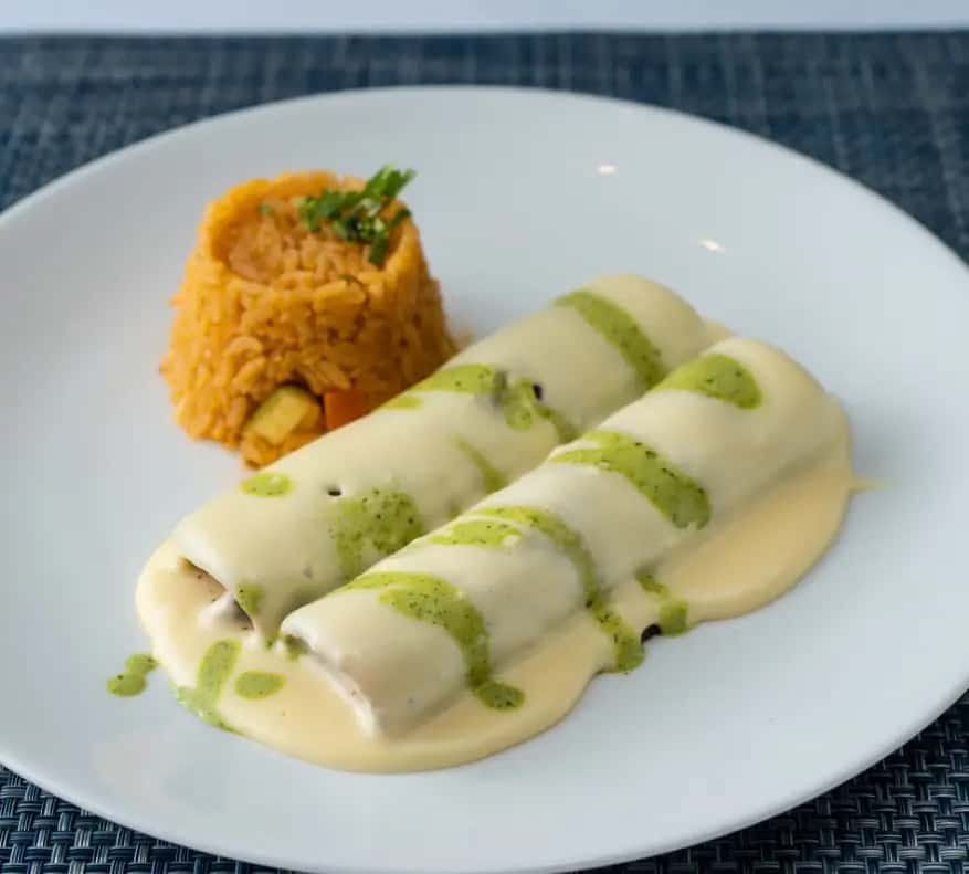 Mesero enchiladas topped with creamy cheese sauce and green drizzle, served with a side of Mexican rice.