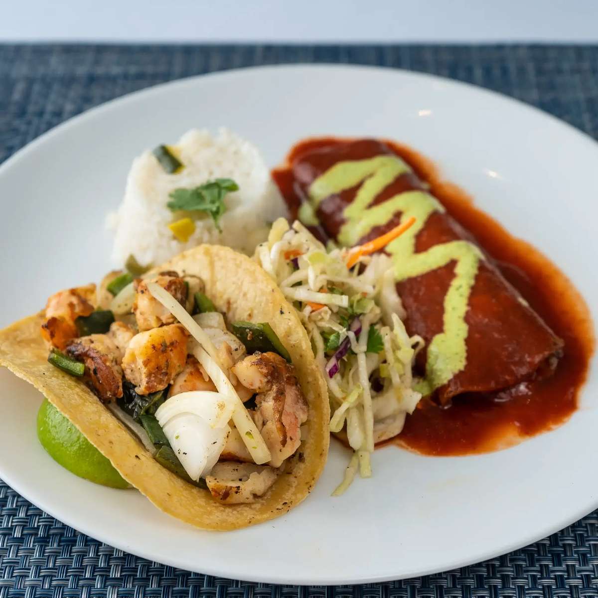 Grilled shrimp taco, enchilada with sauce, rice, and slaw served at Mesero on a white plate.