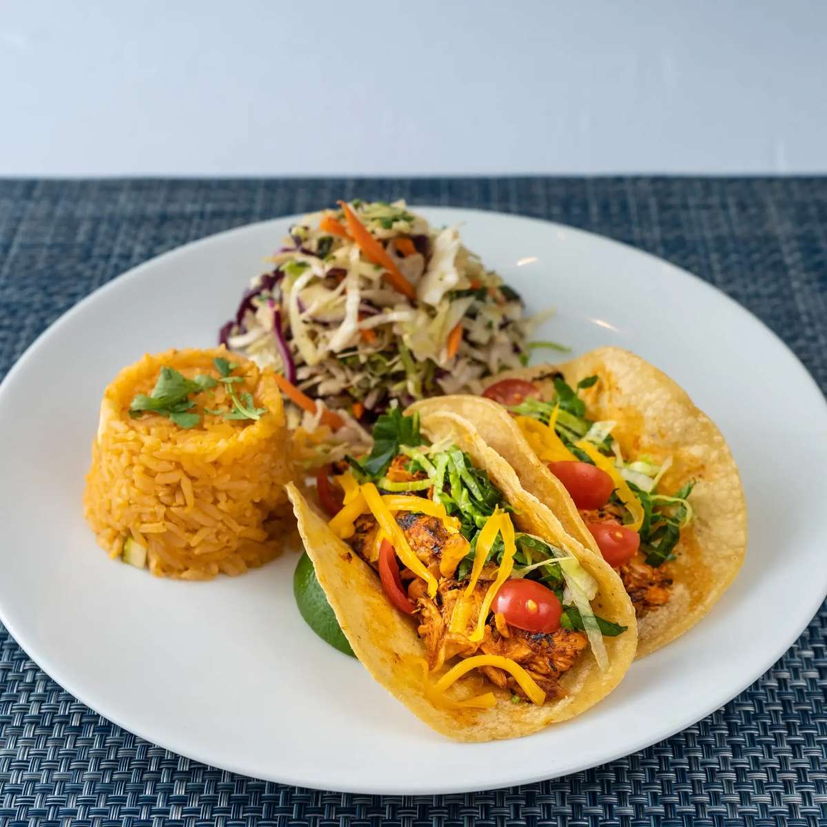 Two corn tacos with shredded lettuce, tomatoes, cheese, and spiced filling, served with yellow rice and cabbage slaw.