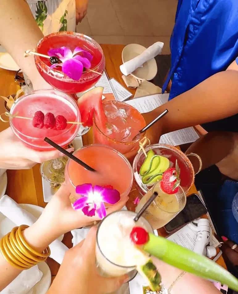 A group of friends raising vibrant craft cocktails garnished with fresh fruit and flowers at Mesero.