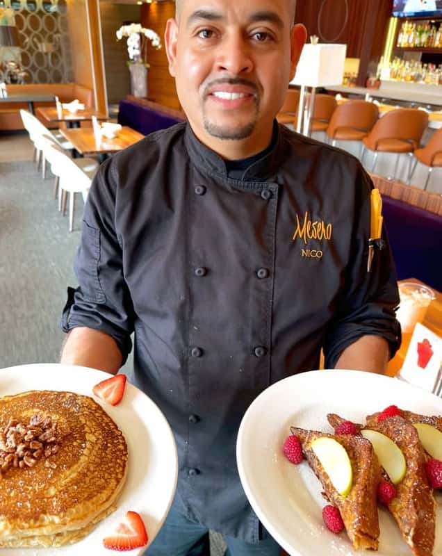 Mesero chef presenting plates of pancakes and French toast topped with fresh fruit inside the warm, modern dining room.