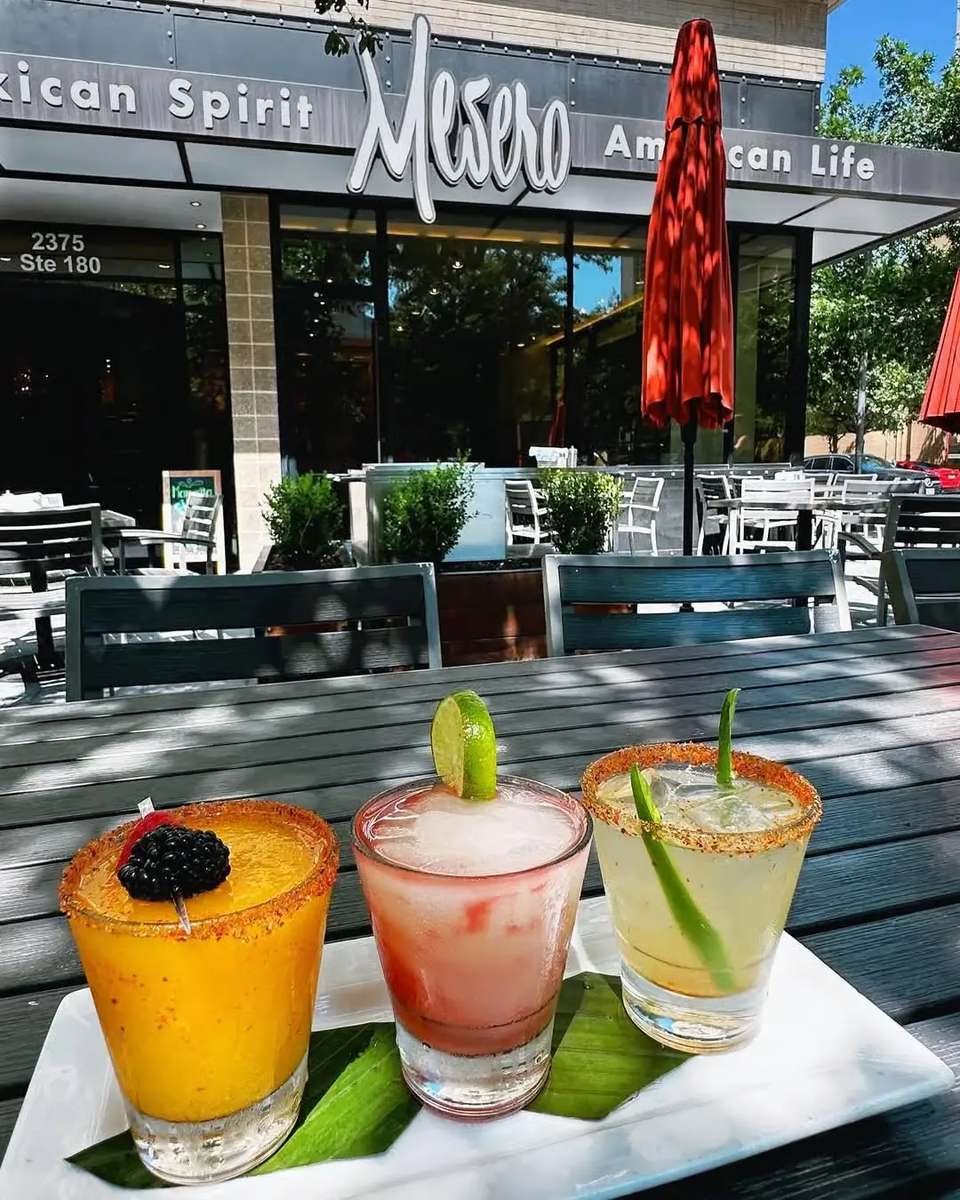 Colorful trio of cocktails on the patio at Mesero, served under red umbrellas with the restaurant sign in view.