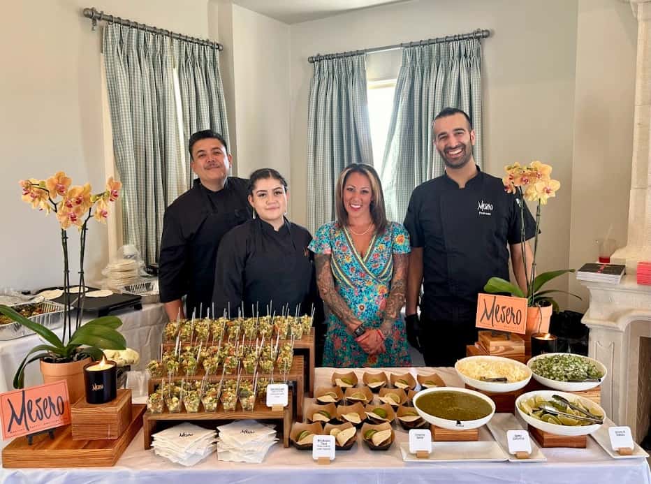 Mesero catering team posing behind the taco bar with fresh salsa, guacamole, and appetizers on display.