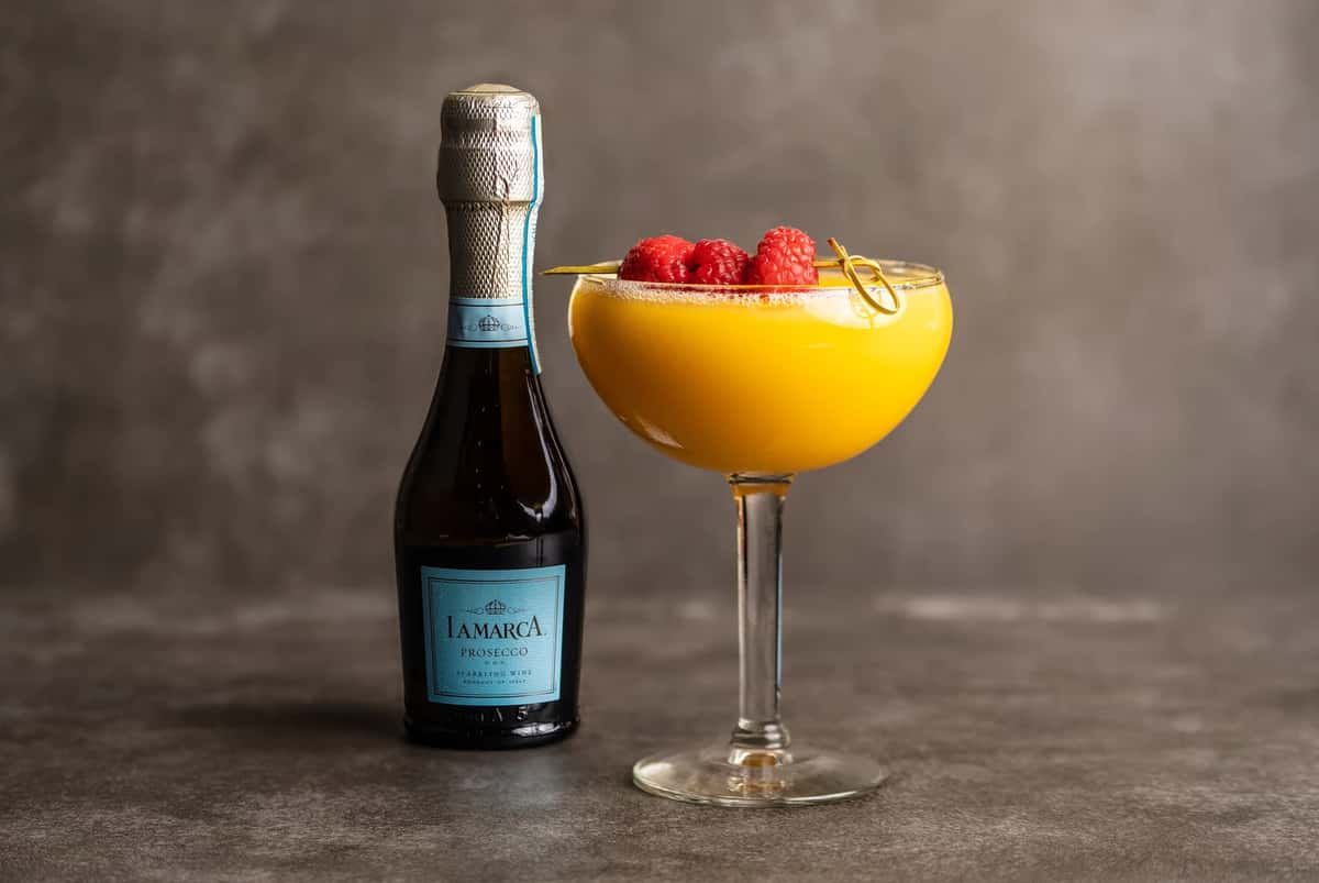 A bottle of La Marca Prosecco next to an orange cocktail in a coupe glass, garnished with fresh raspberries at Mesero.