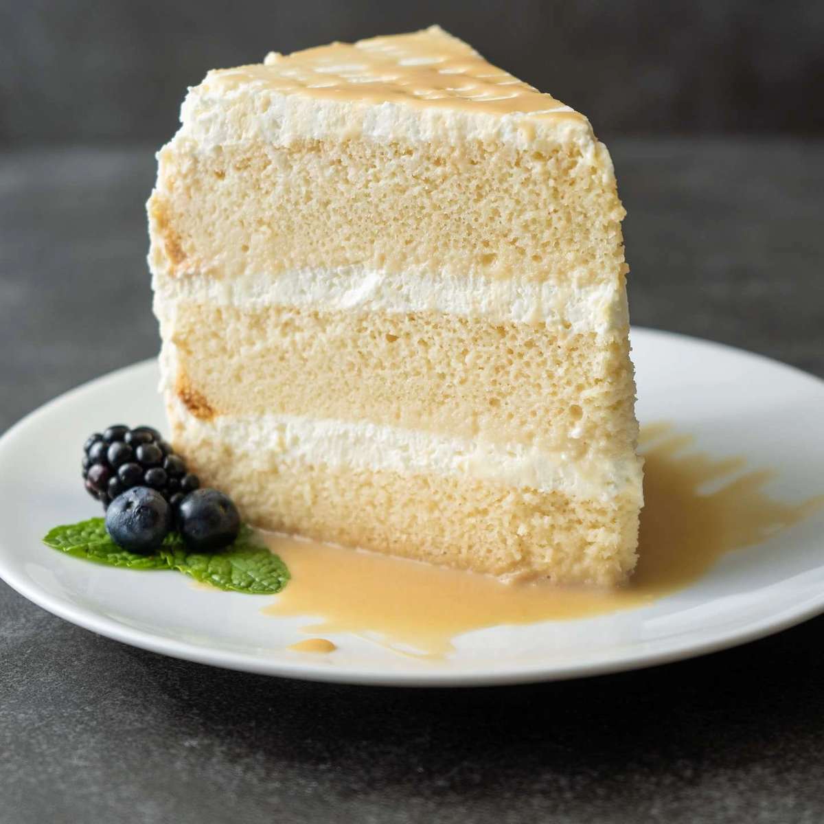 Layered cake slice with white frosting and caramel drizzle, garnished with berries and mint on a white plate at Mesero.