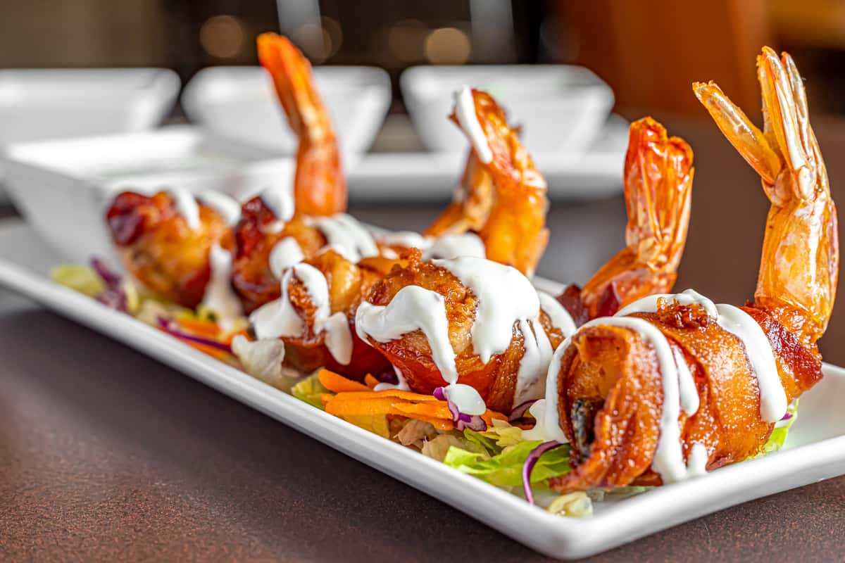  Mesero Bacon Wrapped Camarón featuring grilled shrimp wrapped in crispy bacon, served with fresh sides and vibrant garnishes on a plated dish