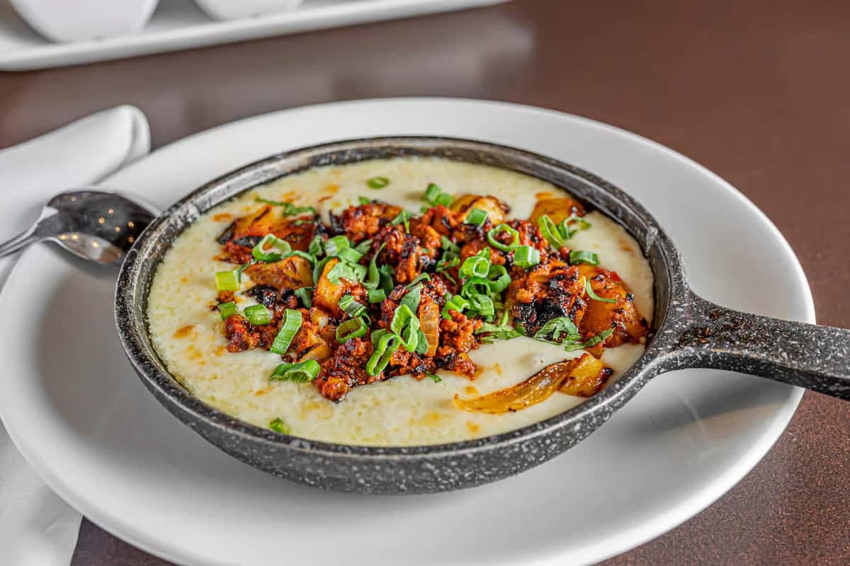 A close-up of a small, rustic skillet of creamy grits topped with chorizo, cilantro, and peppers served at Mesero.