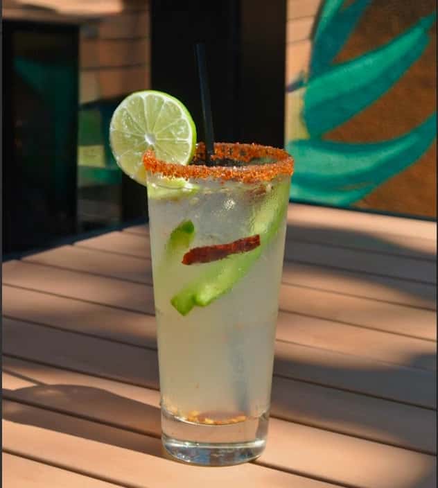 Margarita with chili-salt rim, lime wheel, cucumber slices, and tajín on a sunlit patio table.